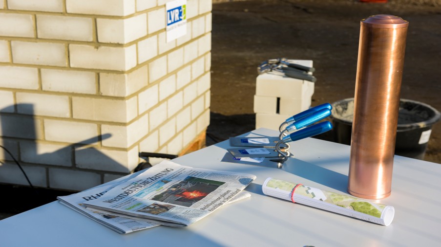 Aktuelle Tageszeitung, Baupläne und Zeitkapsel liegen zusammen mit Maurerkellen auf einen Tisch